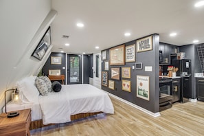 A modern bedroom with a bed, bedside table, and a nightstand with a lamp. There is a kitchen area with appliances and cabinets.