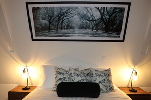 A neatly made bed with two pillows, a black throw, and two bedside lamps. Above the bed, there is a framed black and white photograph of a snowy forest path.