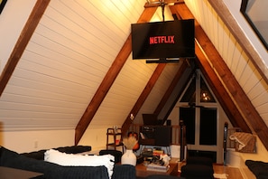 A cozy attic room with a sloped ceiling, a television mounted on the wall, and a small table with various items.