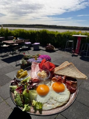 A breakfast tray with eggs, bacon, ham, toast, and salad on a patio with a view of a body of water. - Boutique Stay (Eberswalde)