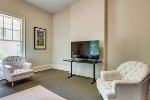 Sala de estar com uma TV de tela grande montada em um suporte, duas poltronas estofadas, um quadro na parede e uma janela com persianas.