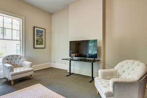 Sala de estar com uma TV de tela grande montada em um suporte, duas poltronas estofadas, um quadro na parede e uma janela com persianas.