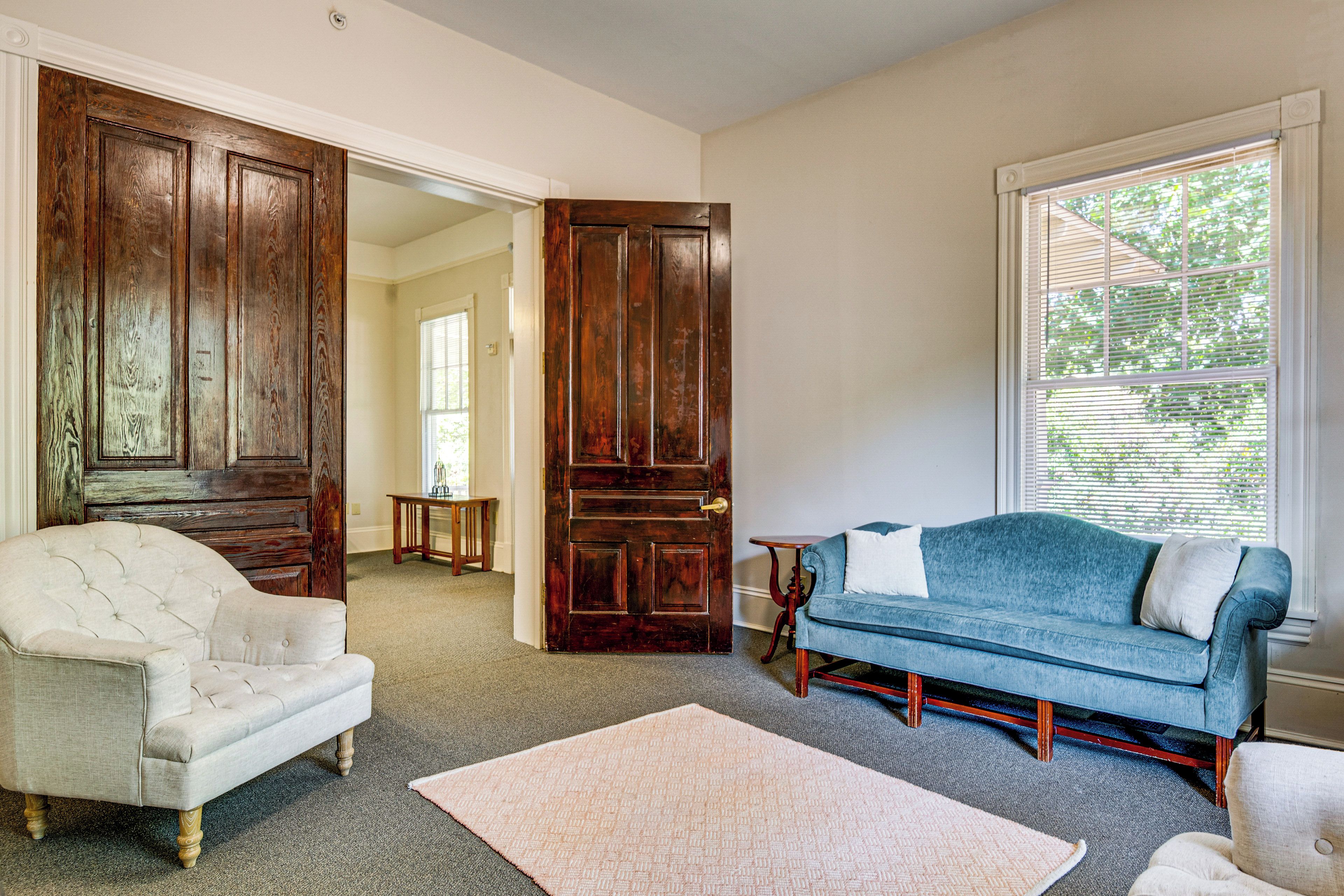 Una sala de estar con un sofá azul, un sillón blanco, una puerta de madera y una ventana con persianas.