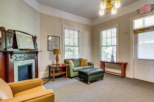 A room with a fireplace, a green sofa, a yellow armchair, a small table with a lamp, and a large window with blinds.