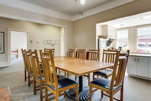 Espaço de refeições com mesa e cadeiras de madeira, cozinha com geladeira e pia, e janela com persianas.