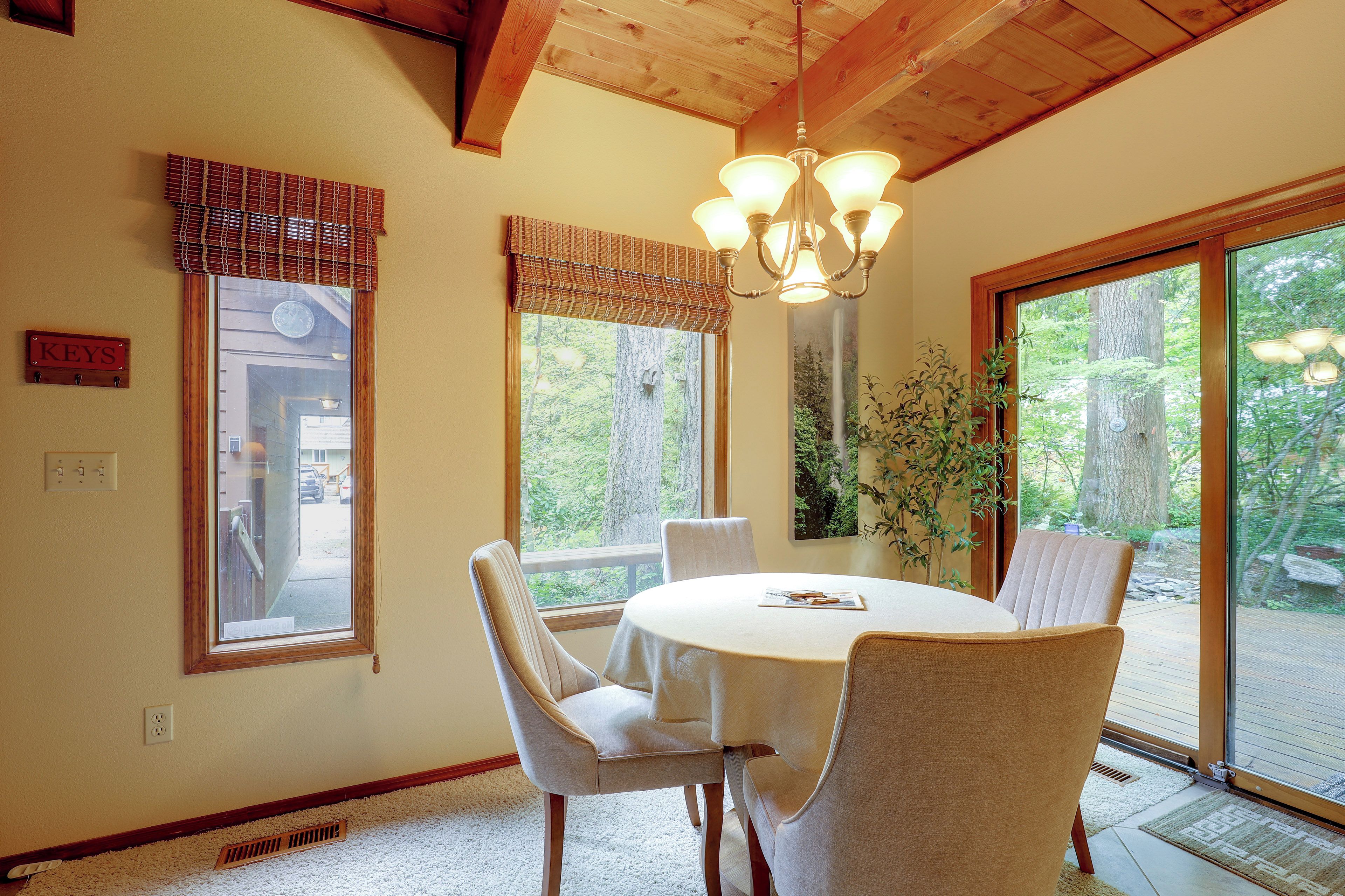 A dining area with a round table, four chairs, a chandelier, and large windows with blinds.