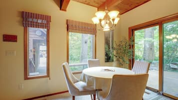 A dining area with a round table, four chairs, a chandelier, and large windows with blinds.