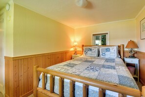 A bedroom with a wooden bed frame, a quilt, a nightstand with a lamp, and a window with a view of trees. - Fire Pit & Views! Lake Champlain Shoreline Stay (Plattsburgh)