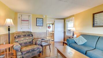 A living room with a sofa, armchair, and a coffee table. There is a door leading to another room, and a framed picture on the wall.