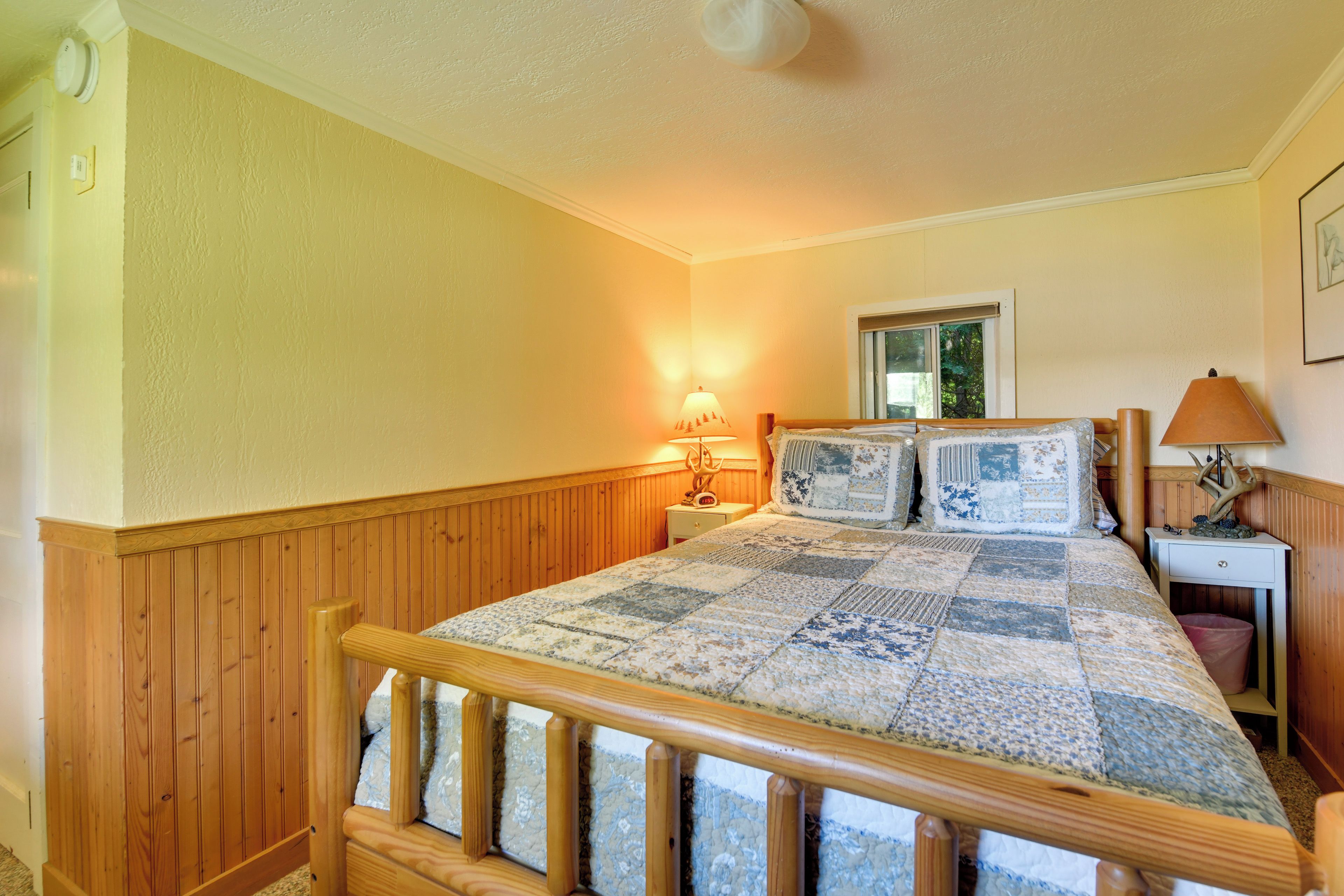 A bedroom with a wooden bed frame, a quilt, a nightstand with a lamp, and a window with a view of trees.