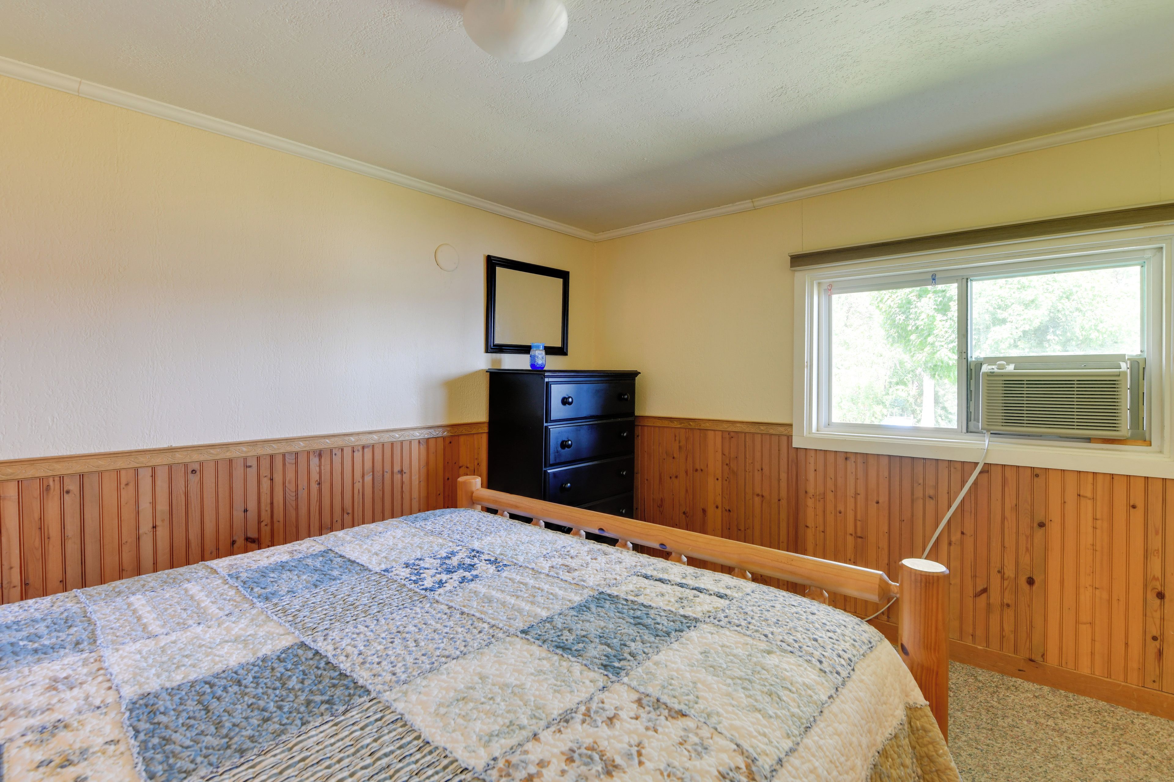 A bedroom with a wooden headboard, a quilted bedspread, a dresser, a mirror, and a window with an air conditioning unit.