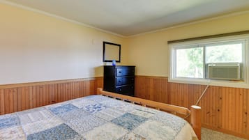 A bedroom with a wooden headboard, a quilted bedspread, a dresser, a mirror, and a window with an air conditioning unit.