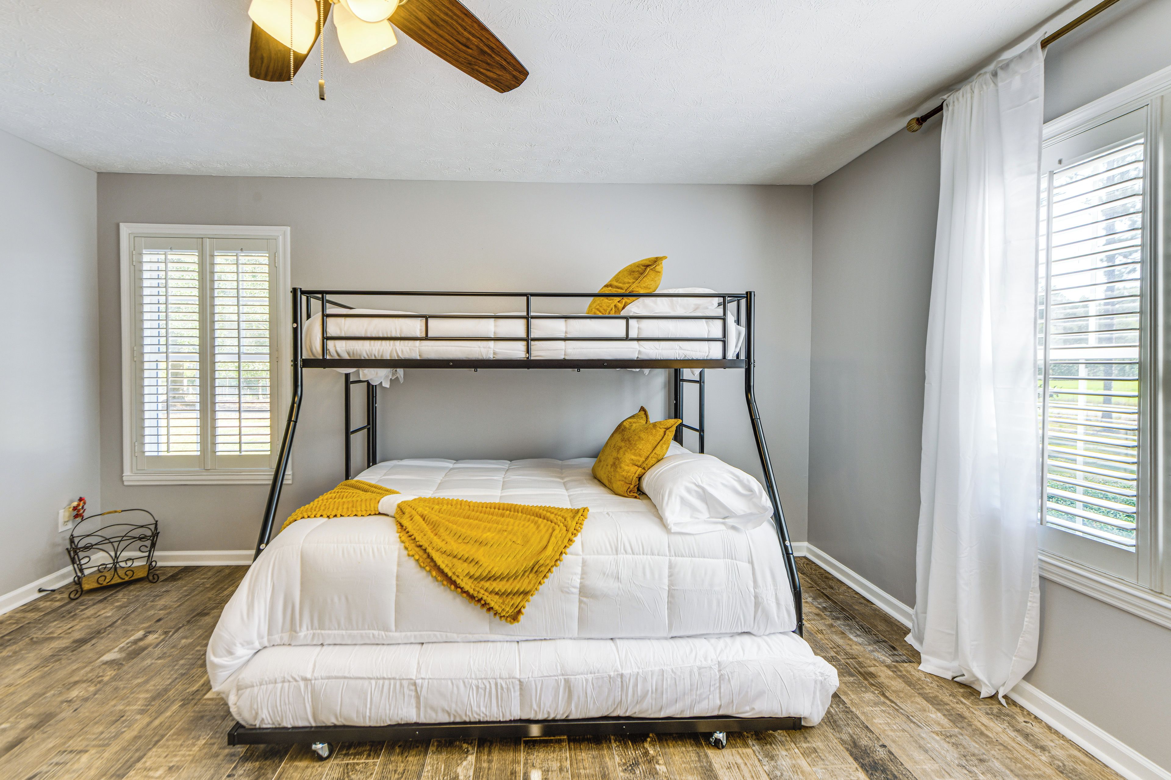A bunk bed with white bedding and a yellow throw blanket, a ceiling fan, and a window with blinds.
