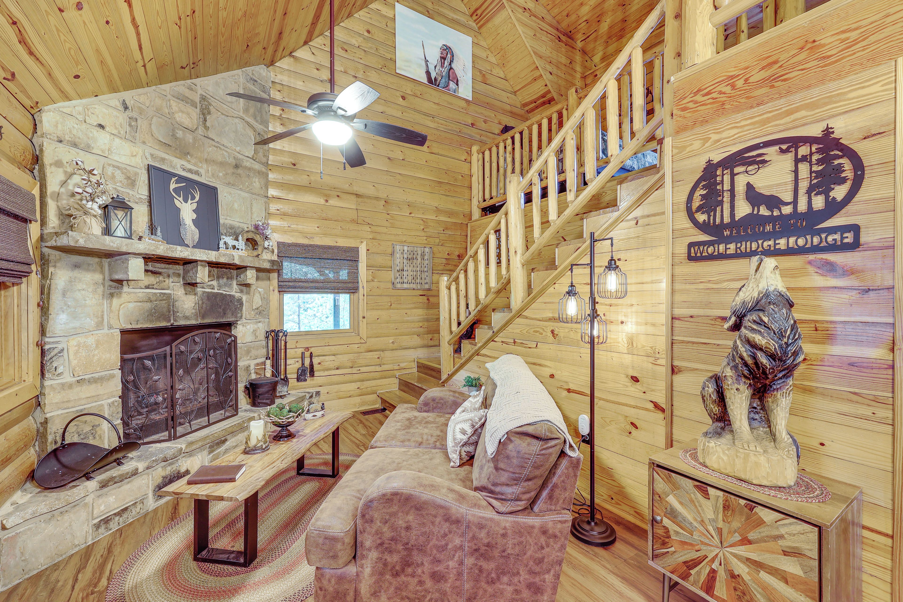 A cozy log cabin interior with a stone fireplace, a wooden staircase, and a welcome sign.