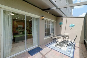 Ein überdachter Patio-Bereich mit einer Schiebetür zu einer Küche, einem kleinen Tisch mit Stühlen und dekorativer Wandkunst mit Palmenmotiven.