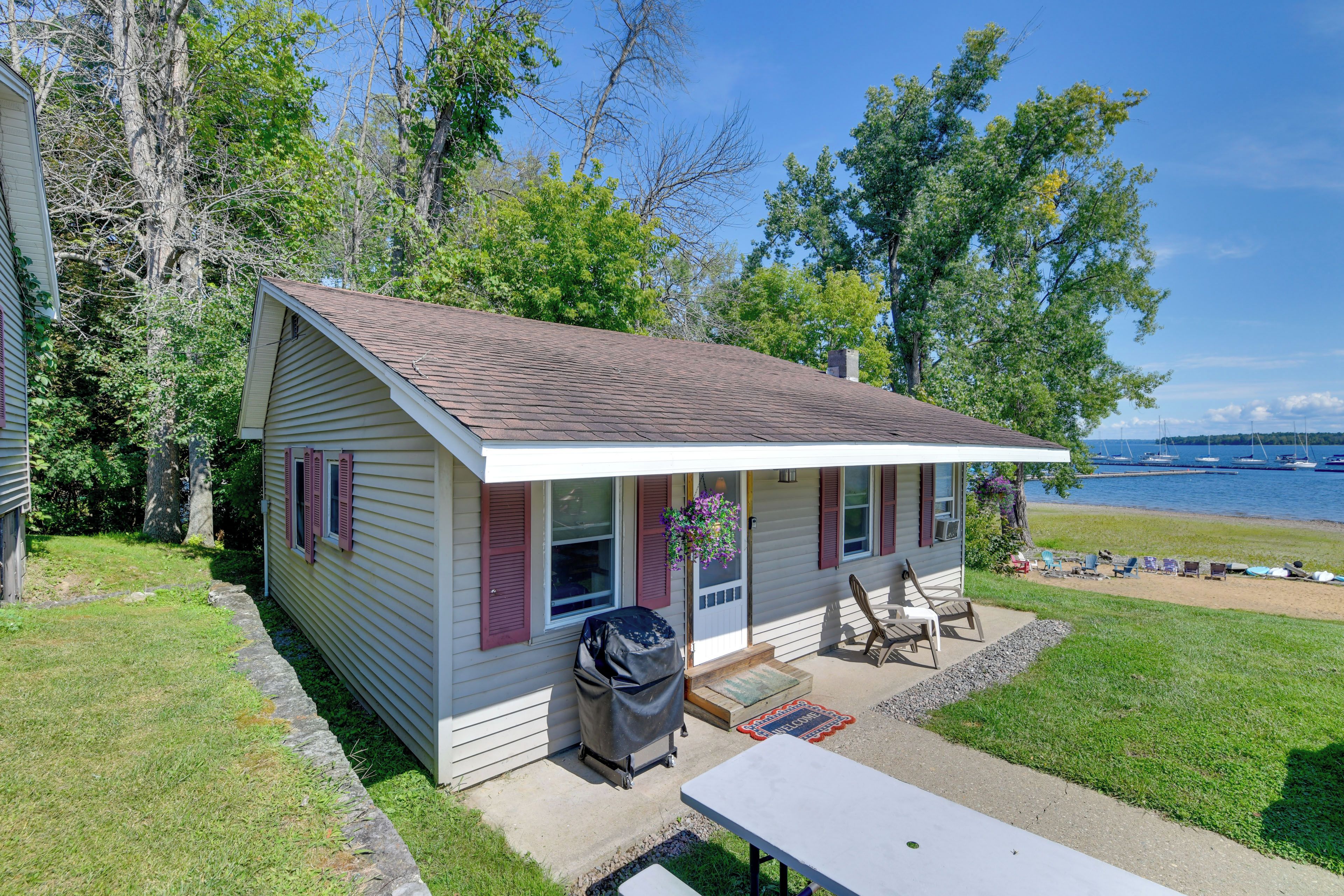A small house with a porch, a table, and a chair, located near a body of water with boats.