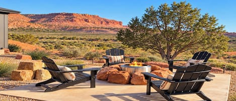 An outdoor seating area with Adirondack chairs and a fire pit, set against a desert landscape with mountains in the background.