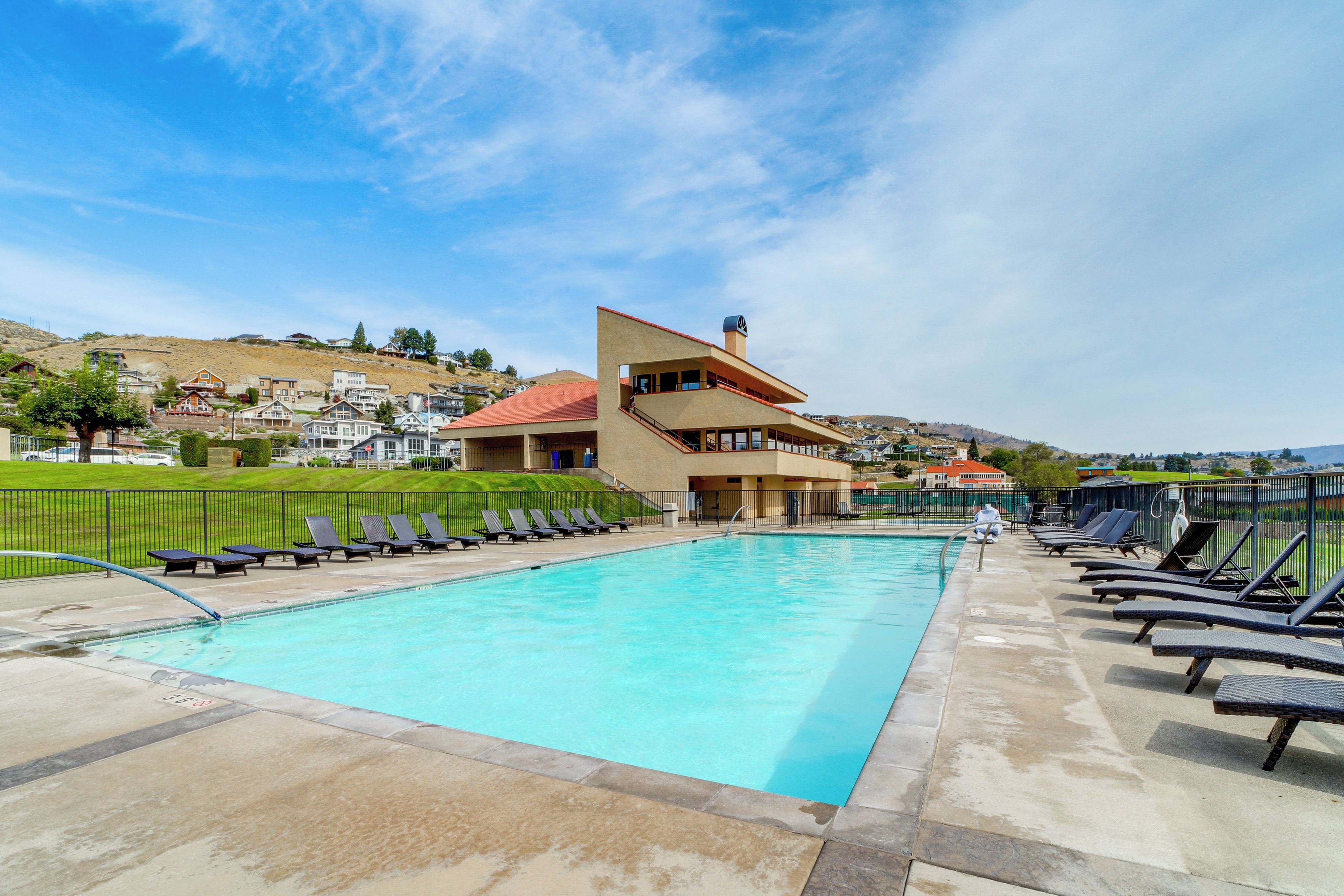 A swimming pool with lounge chairs, a building with a red roof, and a hilly landscape in the background.