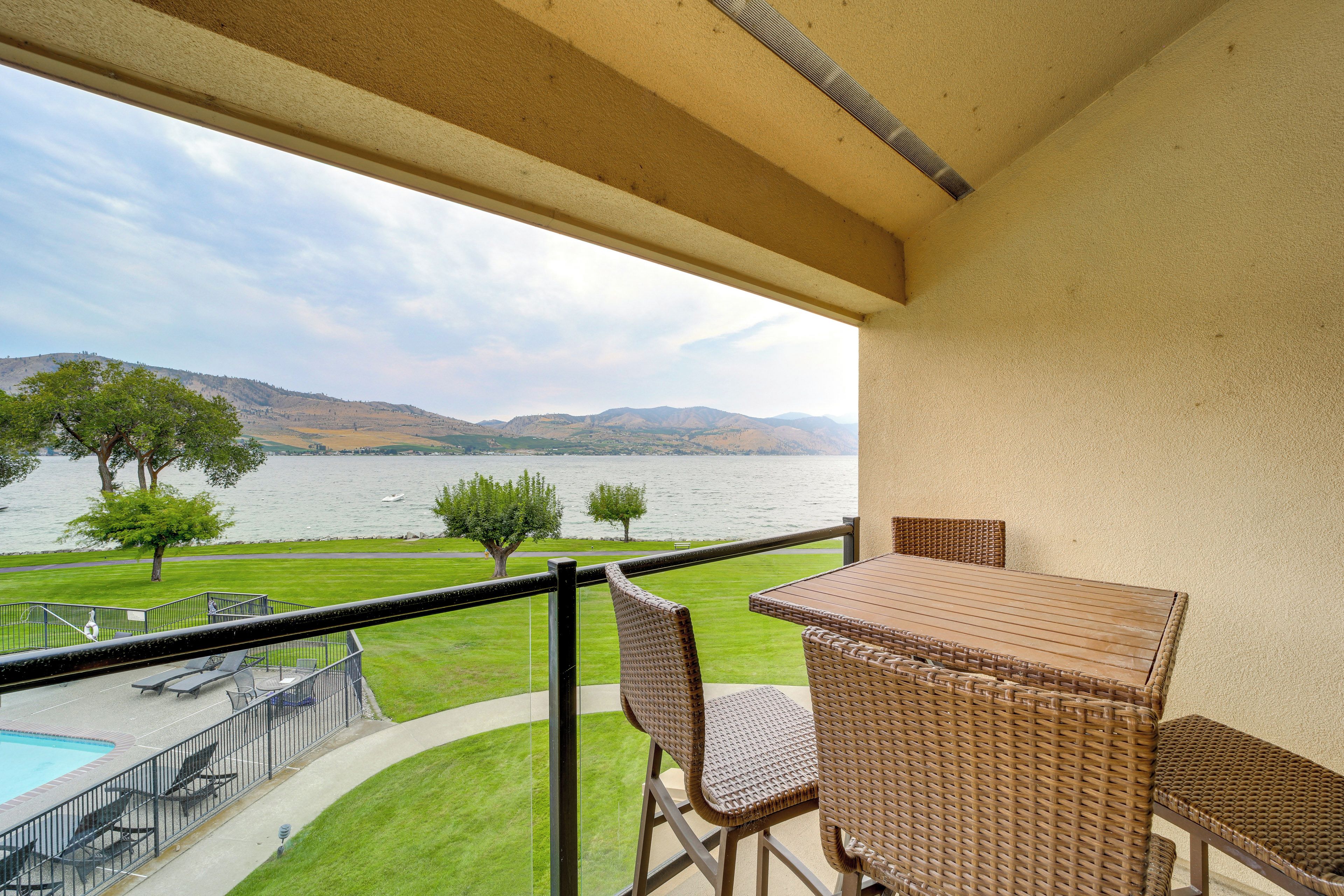 A balcony with a view of a lake and mountains, furnished with wicker chairs and a table.