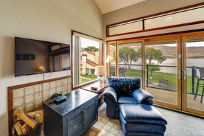 Habitación con una televisión de pantalla plana en la pared, un sofá de cuero, una mesa de madera con una lámpara y vistas a un lago a través de amplios ventanales corredizos.
