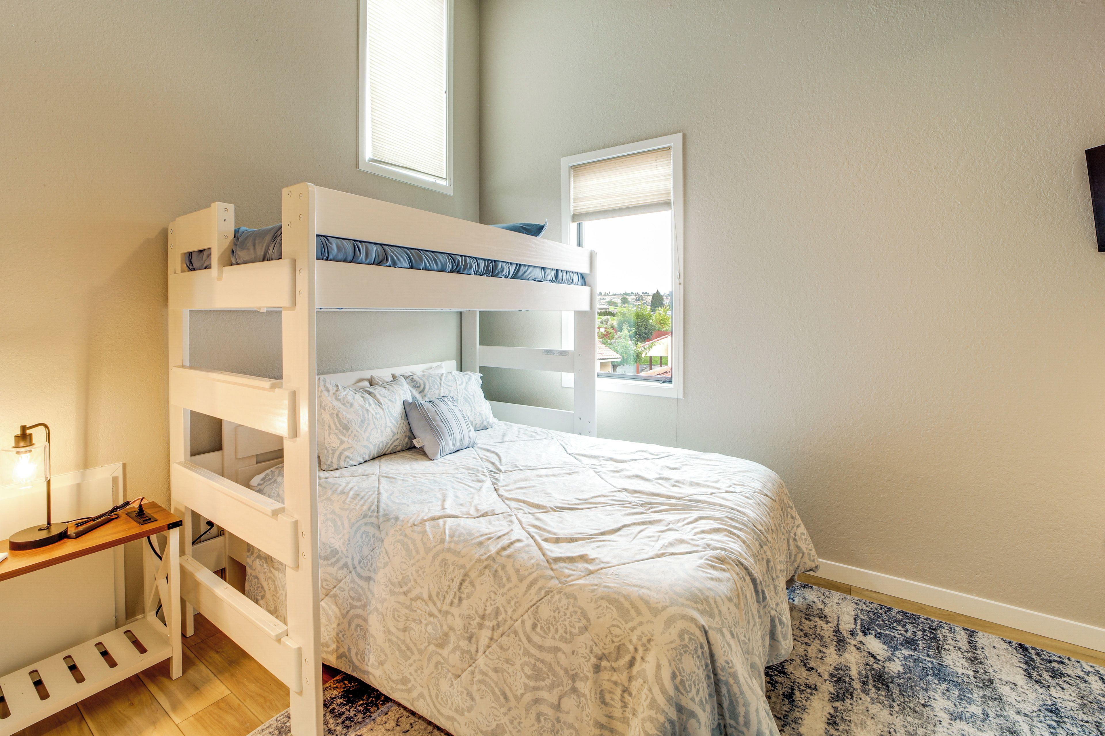 Uma cama beliche com escrivaninha e cadeira, uma luminária de cabeceira e vista para o exterior pela janela.