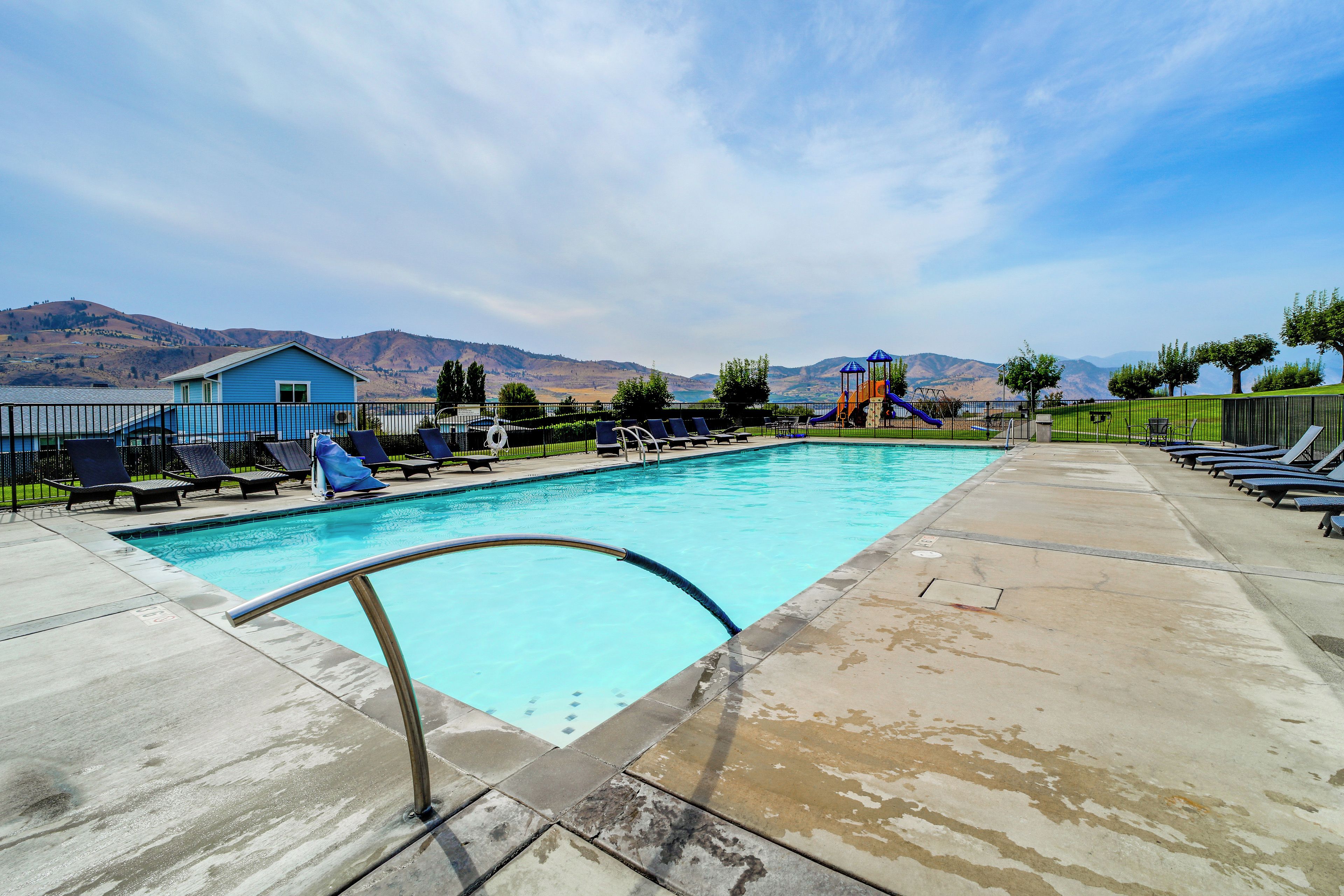 A swimming pool with a metal ladder, lounge chairs, and a playground with a slide.