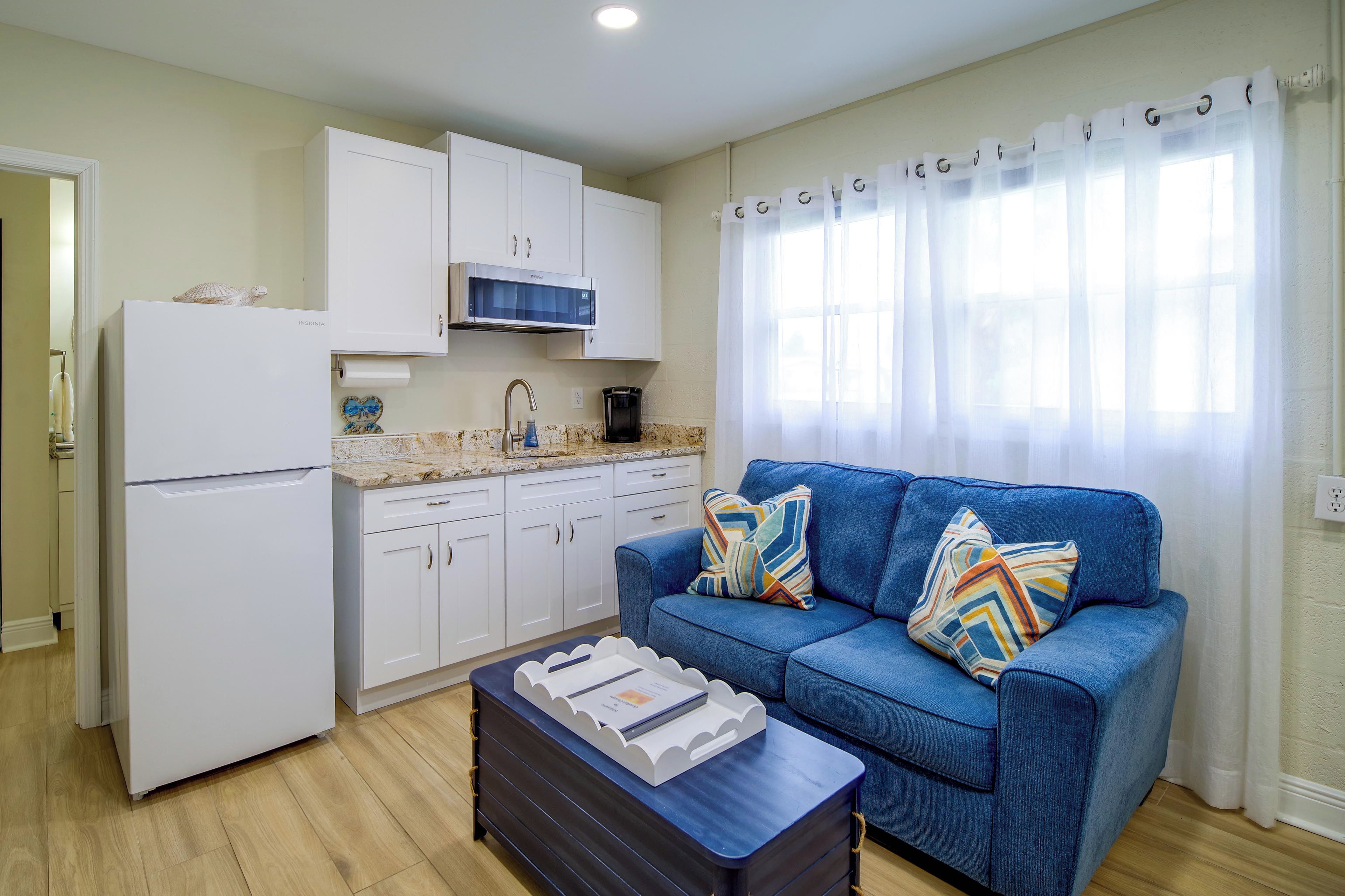 Un espacio habitable compacto con kitchenette, un sofá azul con cojines estampados y una pequeña mesa con una bandeja.