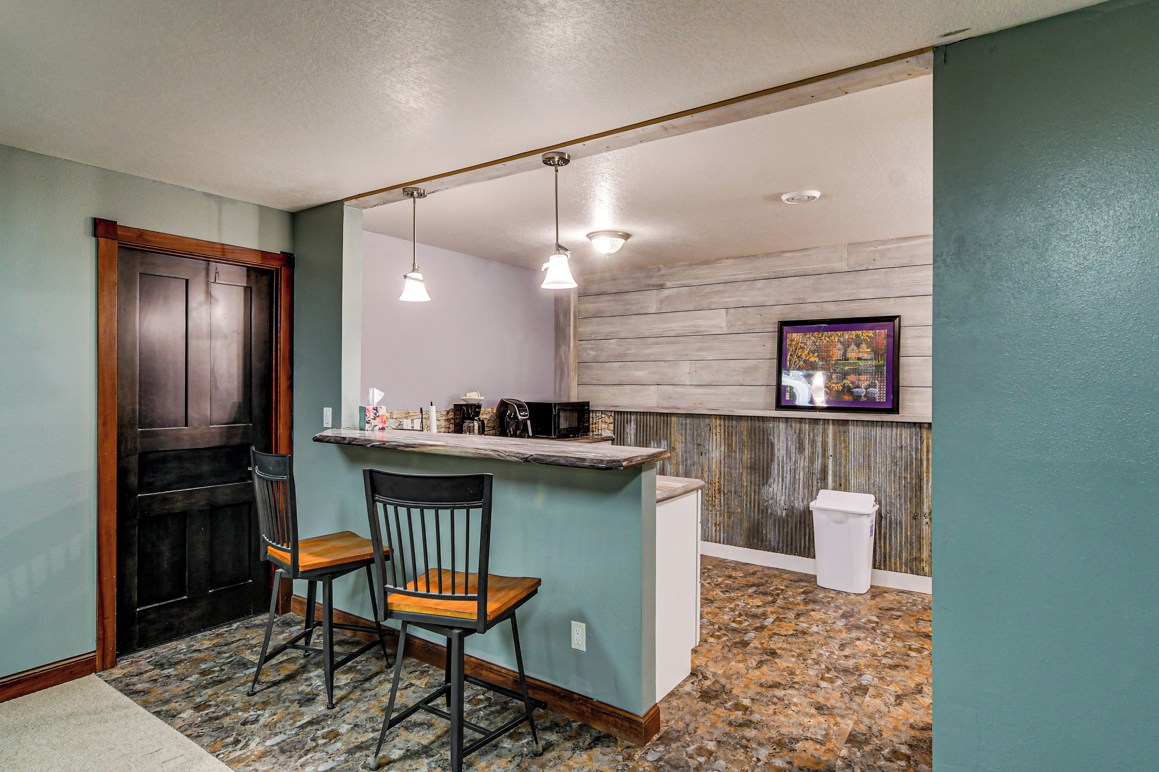 A kitchen with a bar area, two chairs, a trash can, and a framed picture on the wall.