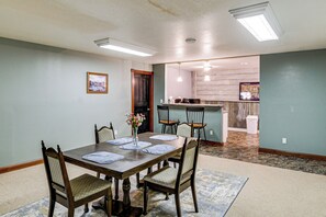 A dining area with a wooden table, four chairs, and a vase of flowers. There is a kitchen in the background with a bar and a trash can.