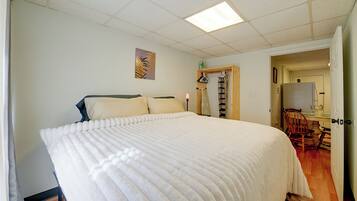 A bedroom with a large bed, a wooden chair, a desk with a chair, and a door leading to another room.