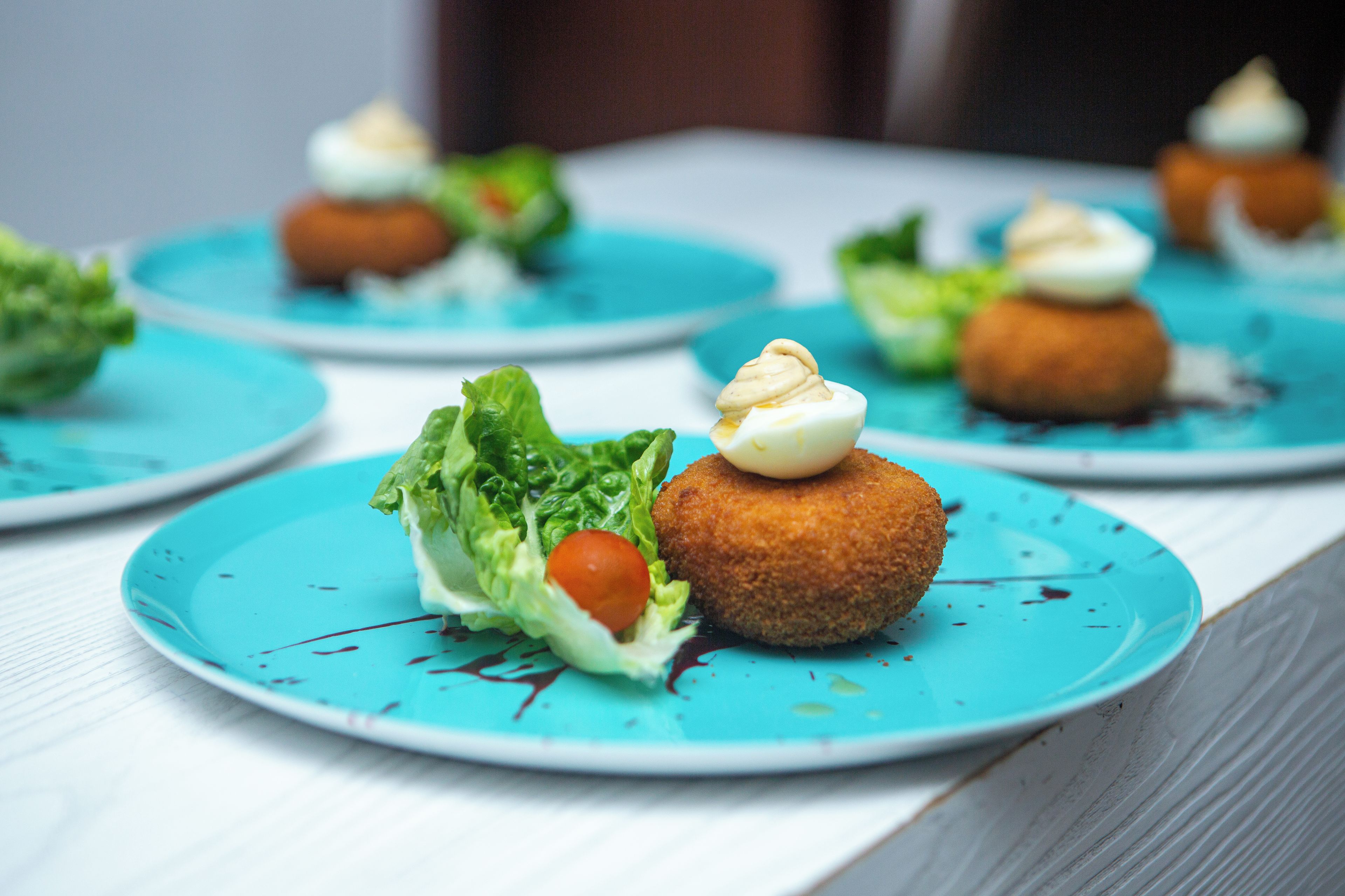 A series of plates with food items, including a round, breaded item topped with a dollop of cream and a small tomato, on a blue plate with a white rim.