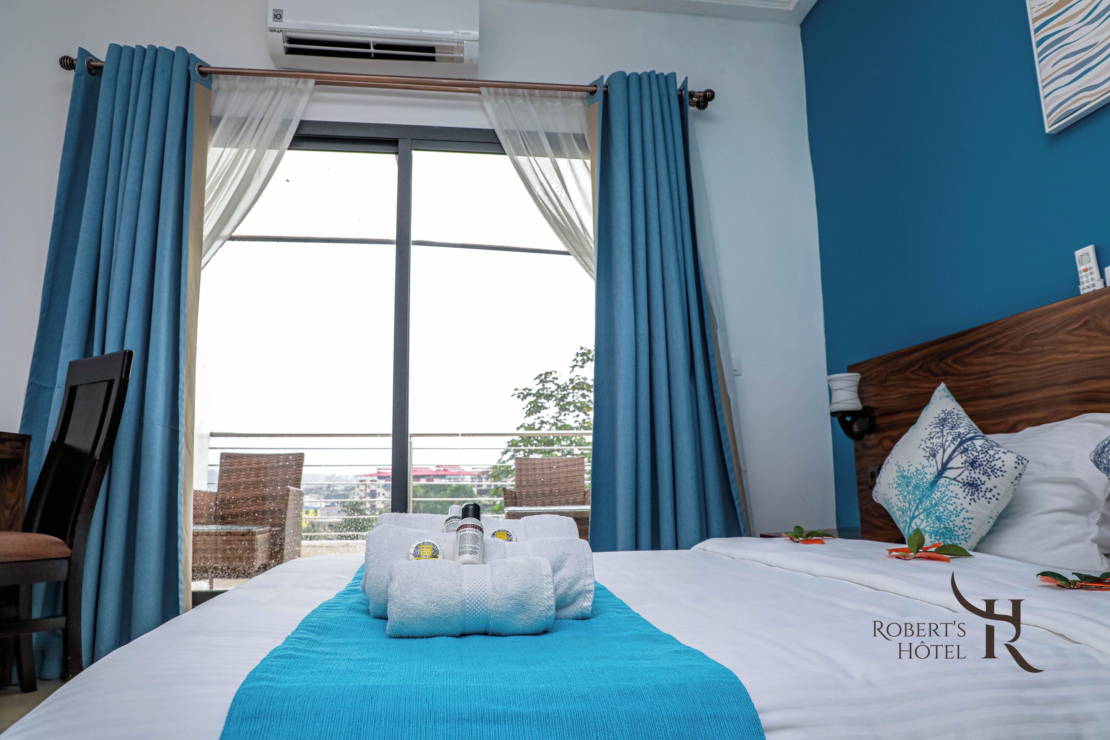 Een hotelkamer met een bed, een raam met gordijnen, uitzicht op buiten, en een bord met de hotelnaam.