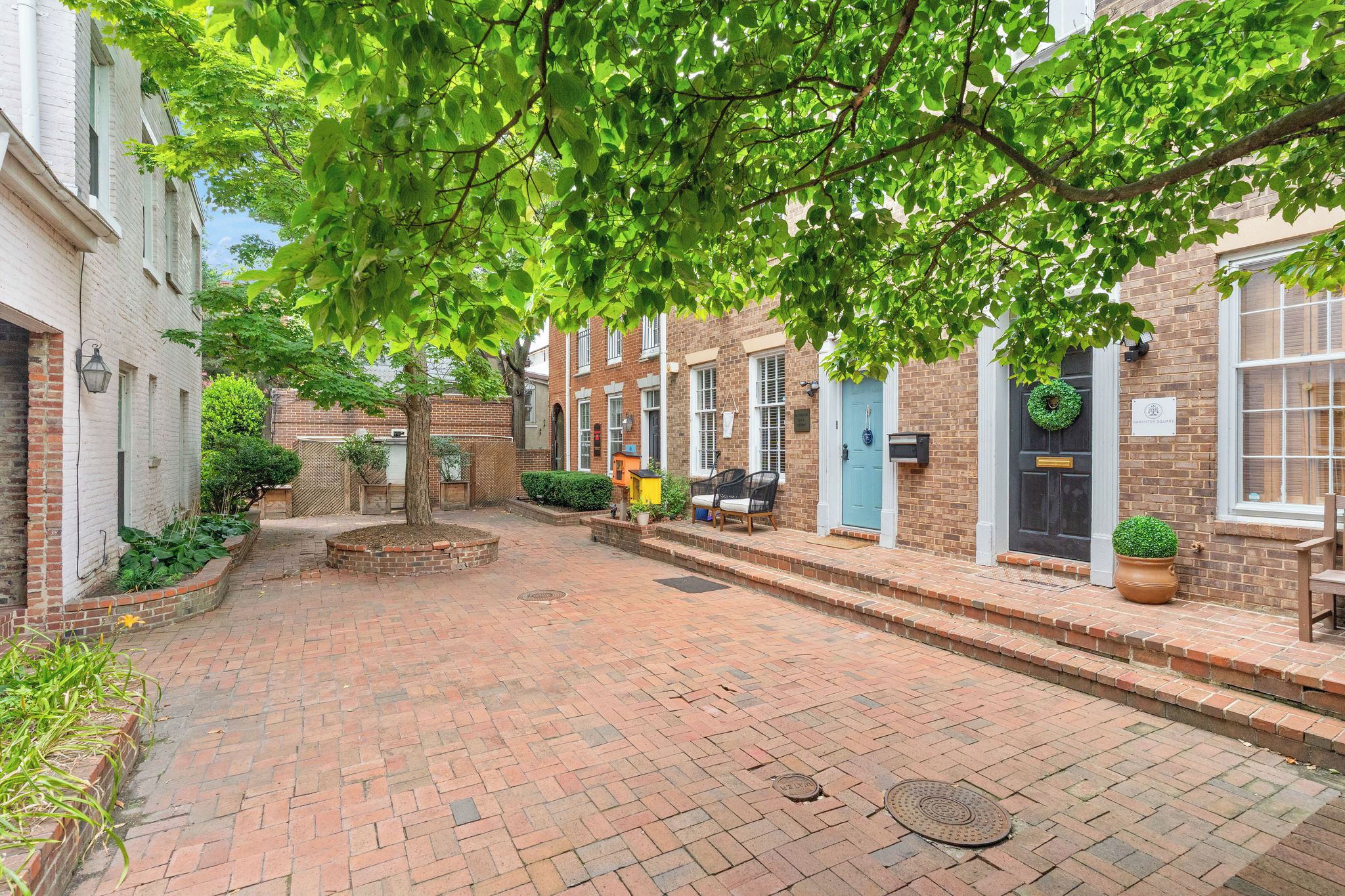 A brick-paved courtyard with a tree providing shade, a bench, and a small garden area.
