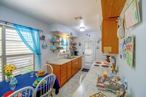 A compact kitchen with a dining area, featuring a small island, wooden cabinets, and a window with blue curtains. - Little Minnow - Just Minutes From the Water (Aransas Pass)