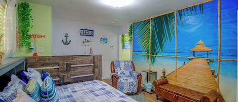 A bedroom with a bed, a wooden dresser, a brown leather sofa, and a large window with a beach mural.