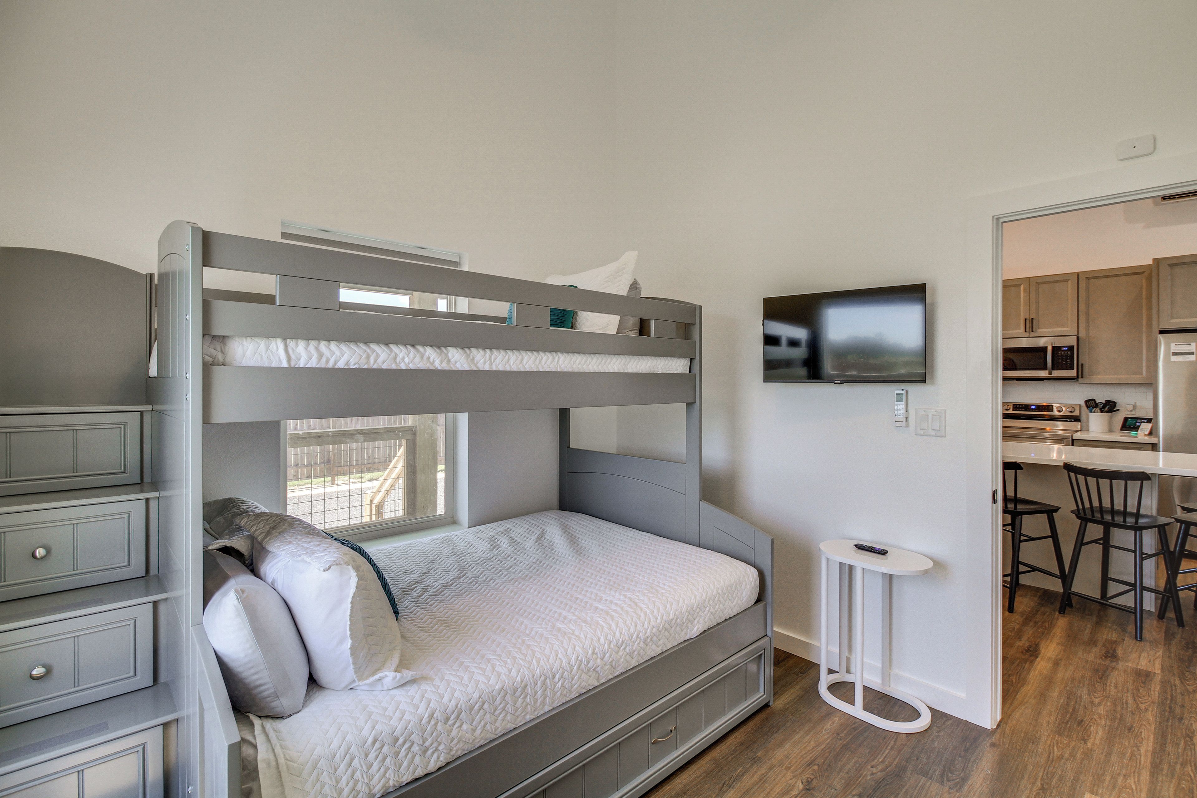 A bunk bed with a desk and drawers, a television on the wall, and a kitchen area with a microwave and stools.