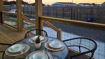 A covered outdoor patio with a table set for four, surrounded by chairs, overlooking a street with a wooden fence and houses.