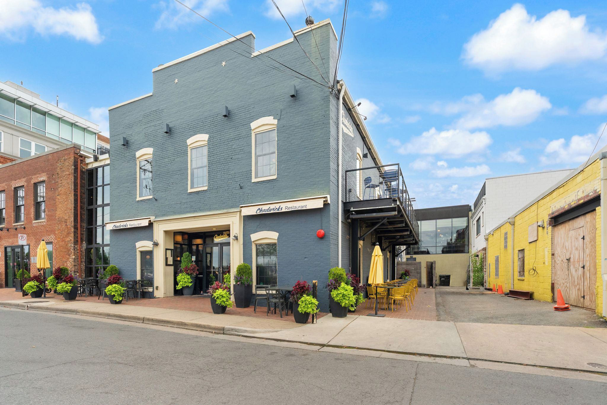 A two-story building with a restaurant named 'Chadwick's Restaurant' on the ground floor. There are outdoor seating areas with tables and chairs, and potted plants are placed around the seating.