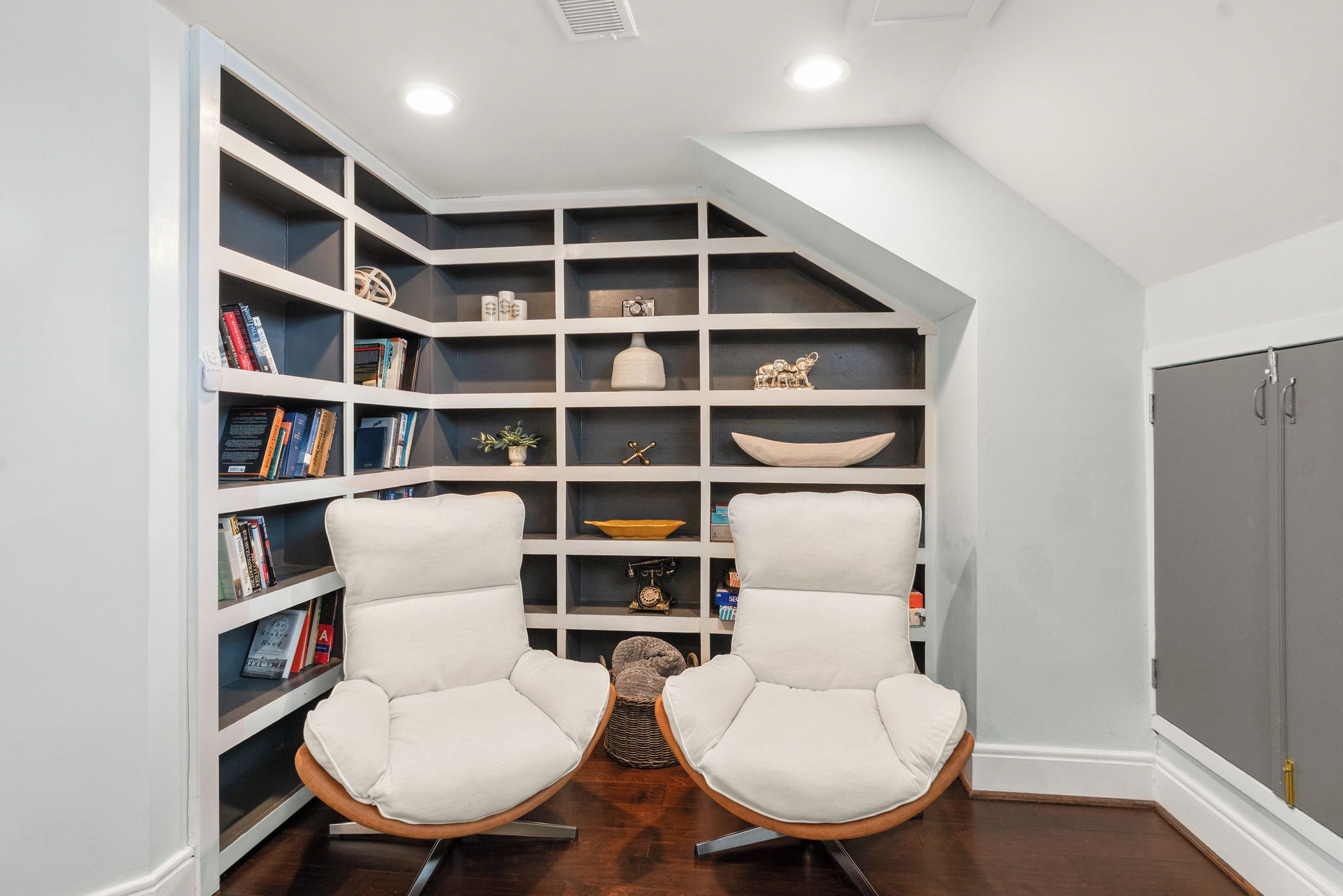 A room with a built-in bookshelf, two white armchairs, a small table, and a grey wardrobe.