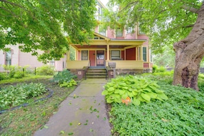 A two-story house with a porch, a front yard with various plants, and a large tree.
