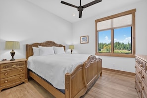 Un dormitorio con una cama de madera, dos mesitas de noche con lámparas, un aparador, un ventilador de techo y una ventana con vistas a árboles.
