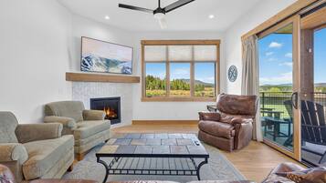 A spacious living room with a fireplace, comfortable seating, a large window with a scenic view, and a ceiling fan.