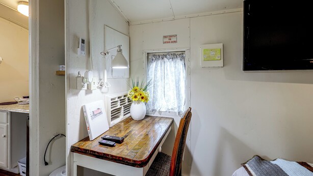 Habitació petita i senzilla amb una taula de fusta, una cadira, un escriptori amb un ordinador, un televisor fixat a la paret i una finestra amb cortines.