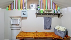 Una cuineta petita amb aire condicionat integrat, una taula de fusta, una estantàcia amb articles de tocador i un ventilador.