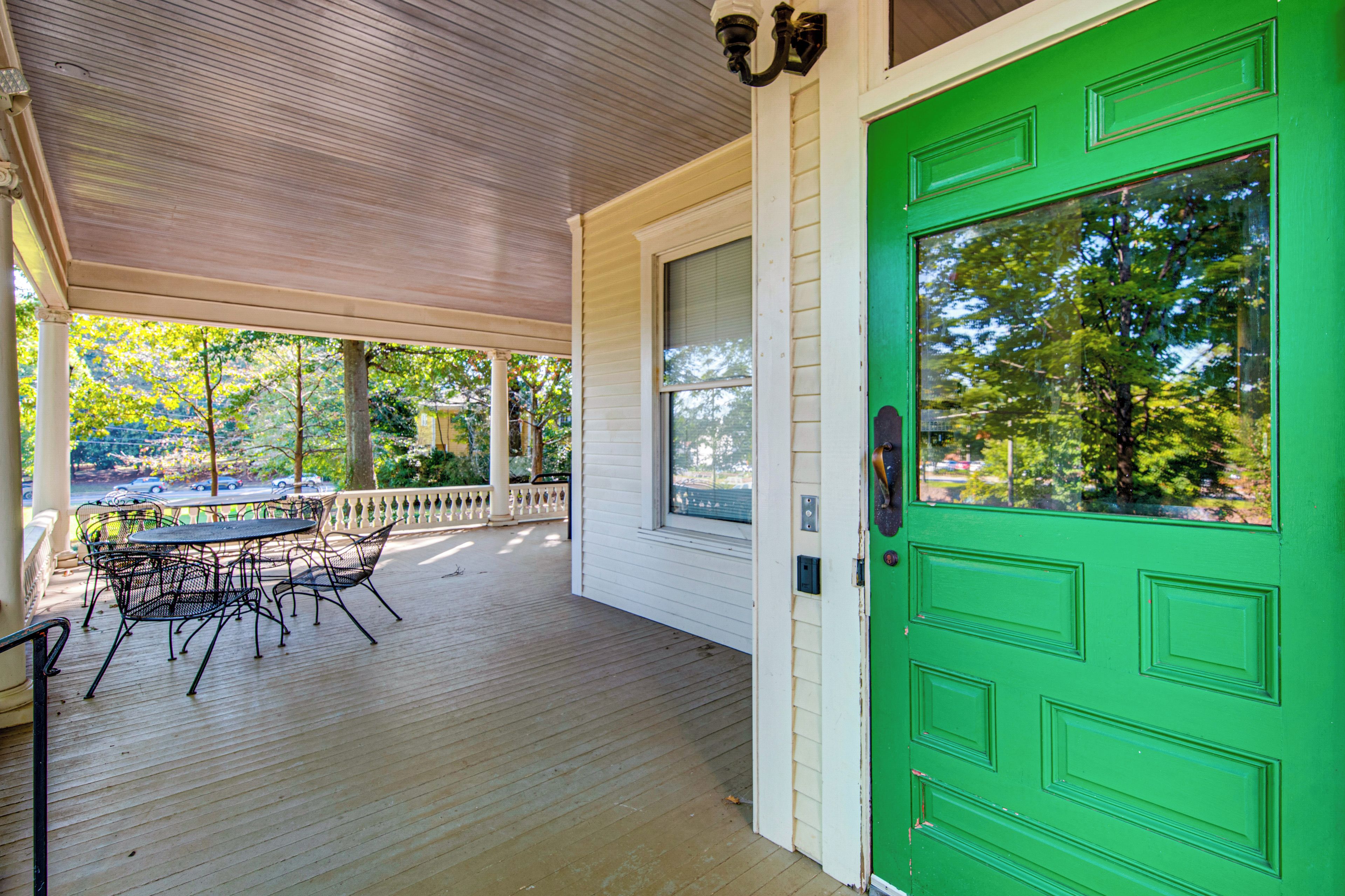 A covered porch with a green door, a table with chairs, and a view of trees and a street.