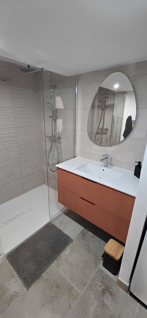 Une salle de bain moderne avec une douche à l’italienne en verre, un lavabo blanc avec un miroir rond et un meuble-lavabo en bois.