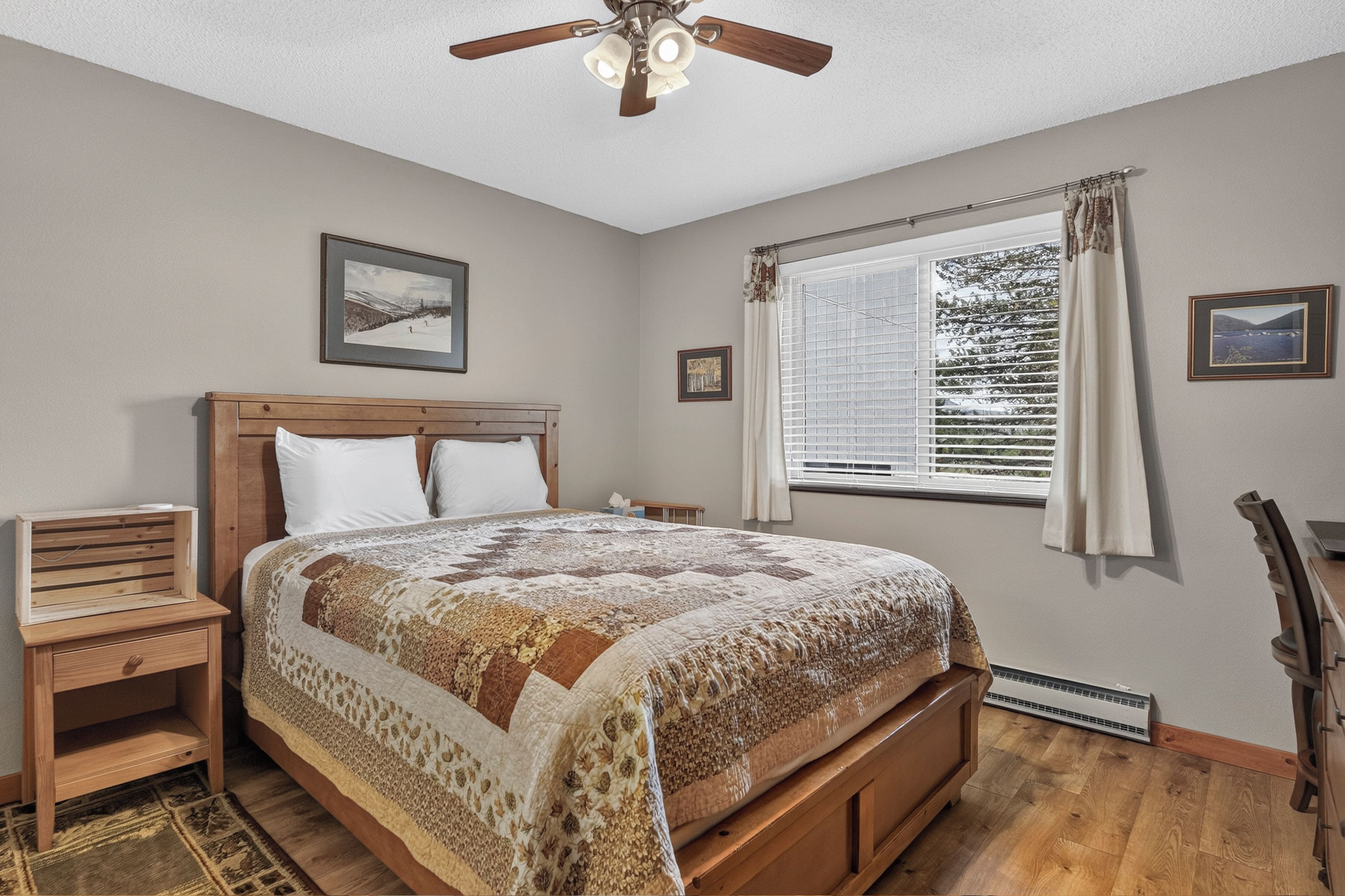 A bedroom with a wooden bed, a nightstand, a ceiling fan, a window with blinds, and framed pictures on the walls.