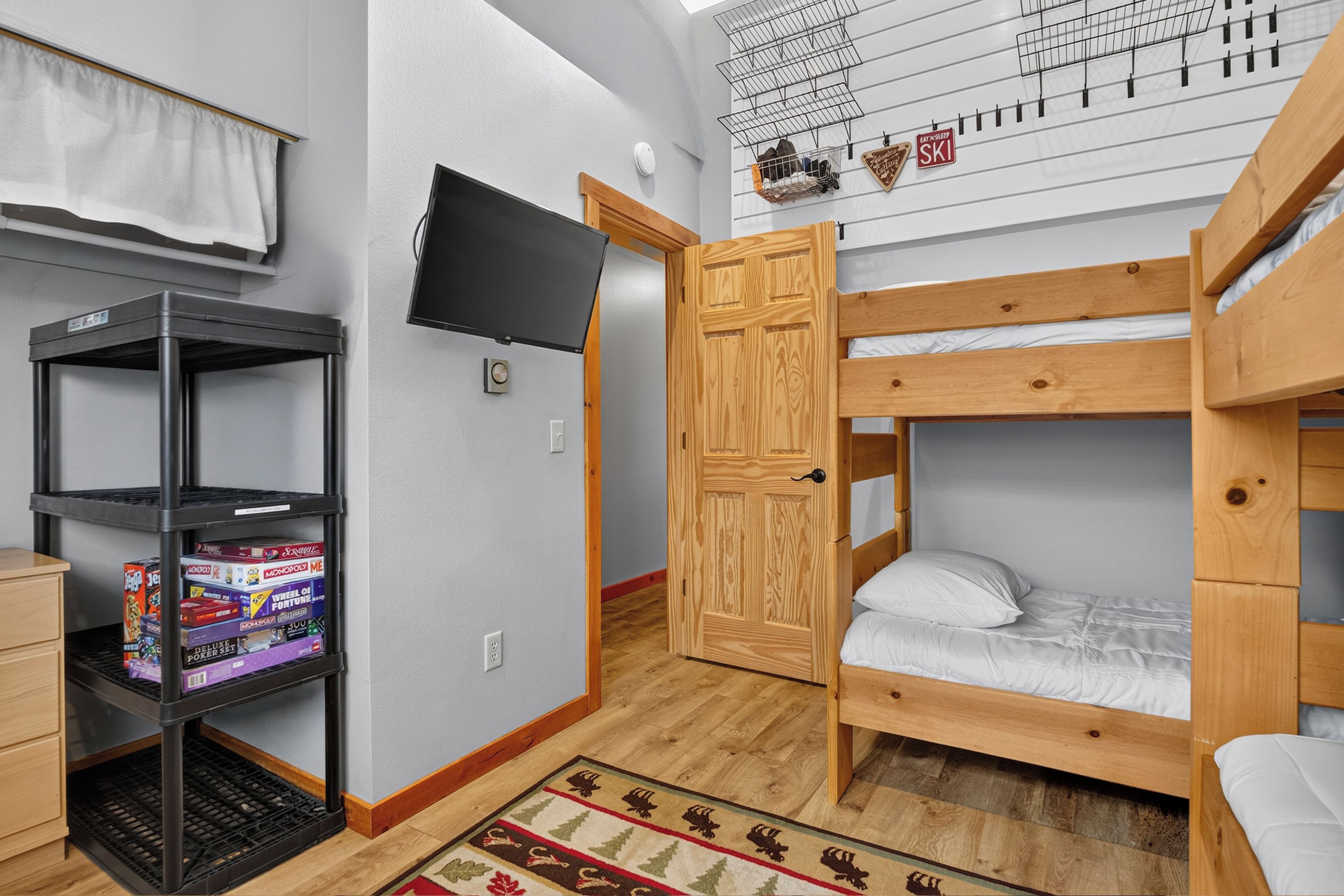 A room with bunk beds, a TV mounted on the wall, a shelf with board games, and a door leading to another room.