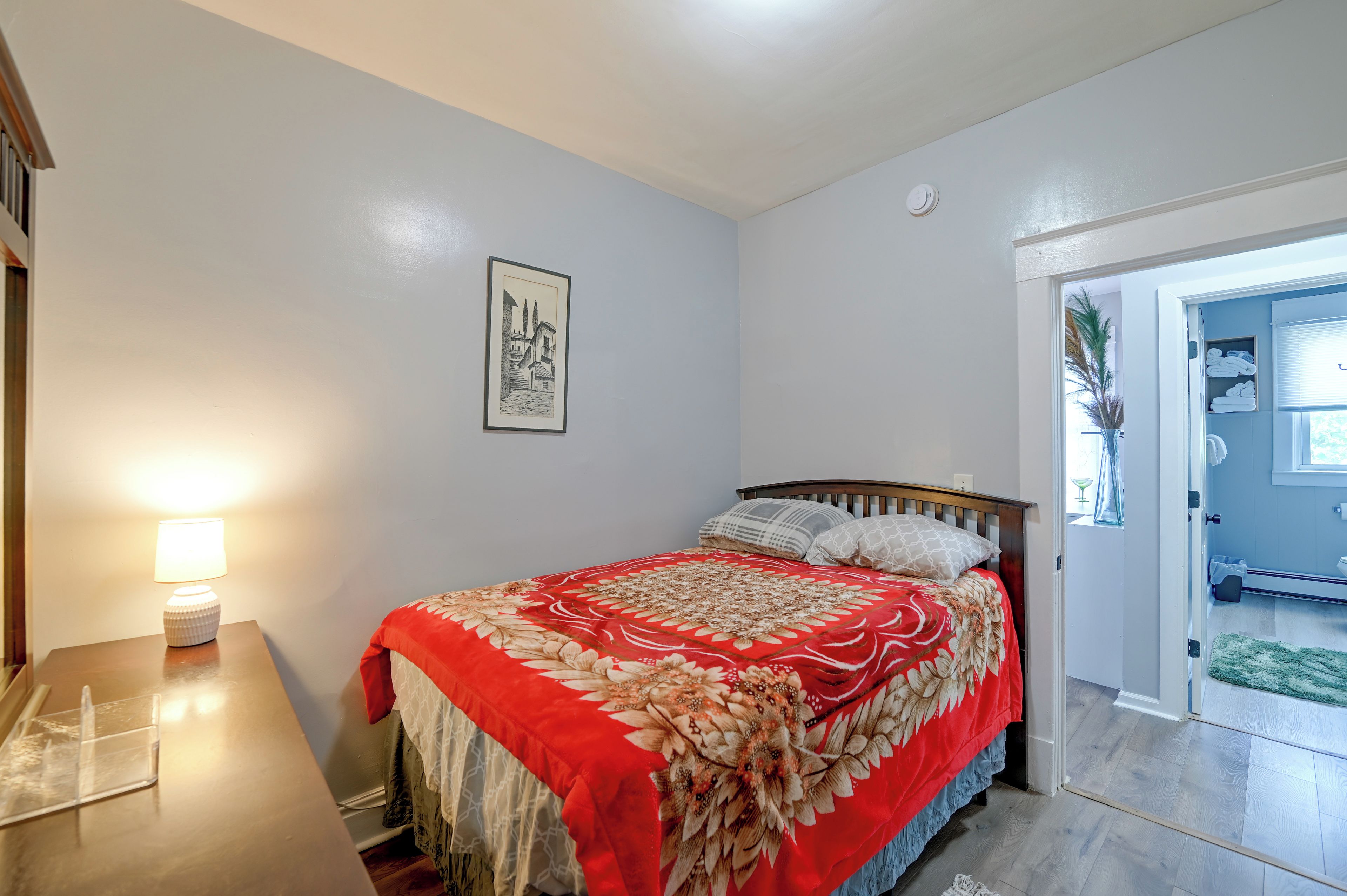 A bedroom with a bed, a bedside table with a lamp, a framed picture on the wall, and a door leading to another room.