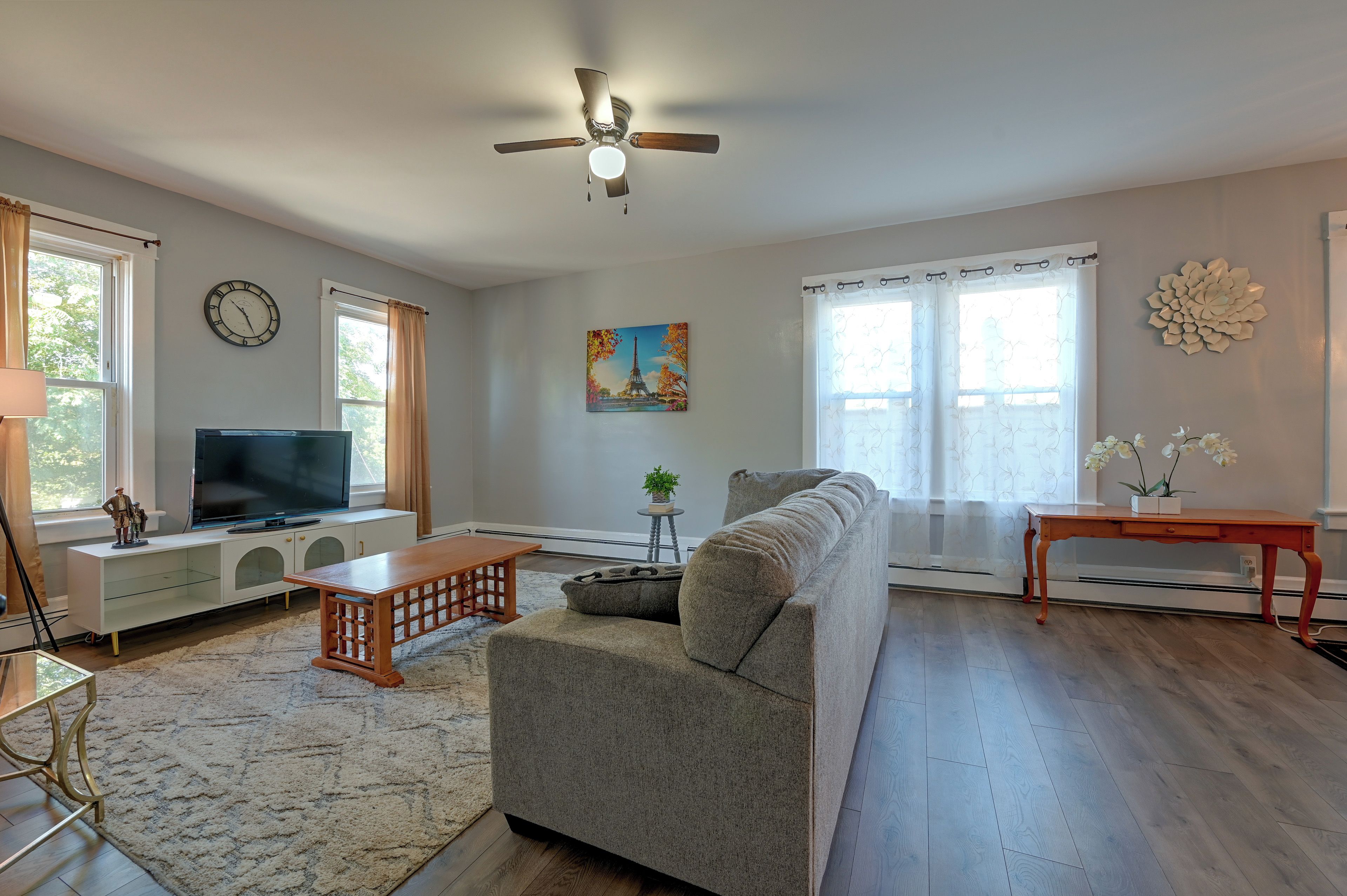 A living room with a sofa, a TV, a ceiling fan, and a painting of the Eiffel Tower.